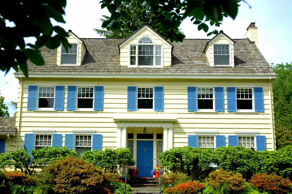 A pale yellow colonial-style house with blue shutters and three dormer windows in the roofline